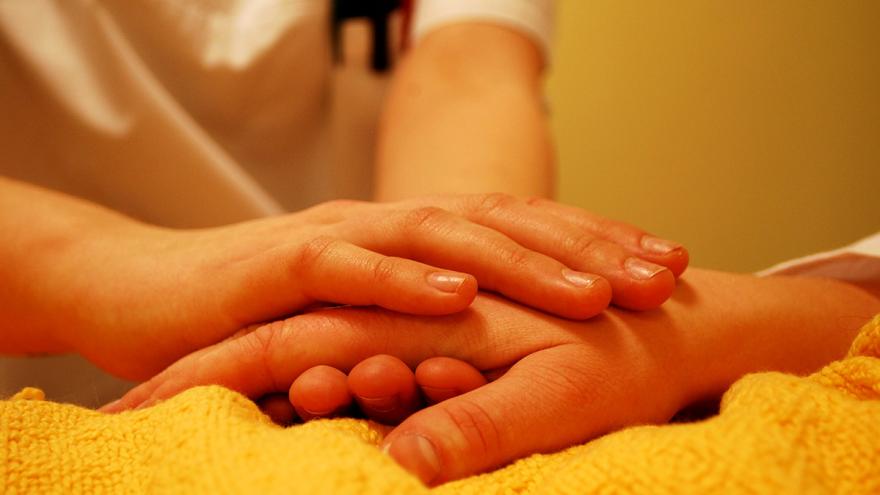 Hände einer pflegenden Person halten die Hand einer im Bett liegenden Person